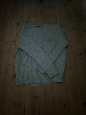 Grå kabelstickad tröja Polo Ralph Lauren - Kabelstickad grå tröja från Polo Ralph Lauren med klassisk rund halsringning och broderad marinblå logga på bröstet. Tröjan har ribbade muddar vid ärmslut och nederkant, och är tillverkad i mjukt material som passar perfekt till höst och vinter.