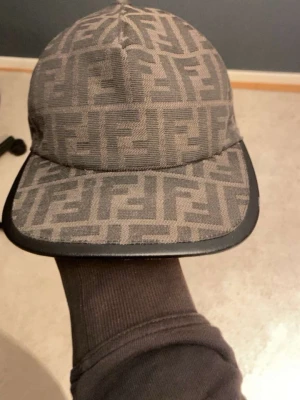 Grå Fendi keps med FF-mönster - Säljer en grå keps från Fendi med det klassiska FF-logomönstret över hela kepsen. Skärmen har en svart kant i läderimitation och baktill finns justerbart band med Fendi-logga. Perfekt för dig som vill ha en exklusiv och trendig accessoar. Priset är ej hugget i sten.