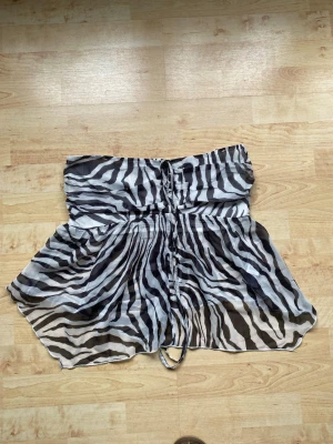 Zebramönstrad bandeau tubtopp - Säljer en zebramönstrad bandeau topp med svartvita ränder. Toppen har resår upptill och nedtill samt snörning framtill. Materialet är tunt och luftigt, perfekt för sommaren. Passar dig som vill sticka ut med ett djurmönster.