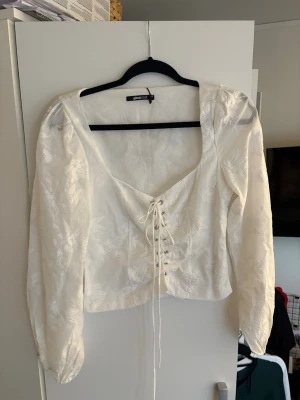 Vit blus med snörning från Gina Tricot - Stilren vit blus från Gina Tricot med långärmad design och vacker blommig struktur. Blusen har djup urringning och snörning framtill som ger en trendig touch. Materialet är lätt och har en subtil glans som gör den extra snygg.