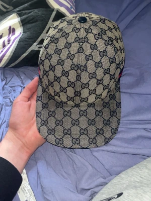 Beige Gucci keps med GG-mönster - Säljer en beige keps från Gucci med klassiskt GG-mönster över hela kepsen. Kepsen har böjd skärm och justerbar rem baktill i mörkt läder. Materialet är canvas med läderdetaljer och kepsen har en cool och lyxig vibe som sticker ut.