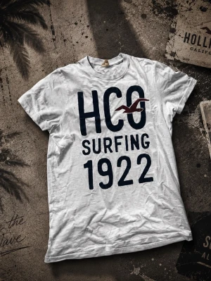 Vit HCO t-shirt med tryck - Vit t-shirt från Hollister med stort mörkblått tryck 'HCO Surfing 1922' och en röd fågel på bröstet. Klassisk rund hals och korta ärmar. Perfekt för dig som gillar surfstil och avslappnad look.