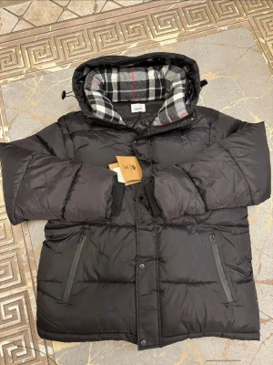 Svart pufferjacka med rutigt foder - Snygg svart pufferjacka från Burberry med klassiskt rutigt foder i svart, vitt och rött. Jackan har huva, dragkedja och tryckknappar framtill samt två snedställda fickor med dragkedja. Perfekt för kalla dagar och riktigt trendig look.
