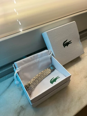 Silverfärgat armband från Lacoste - Snyggt armband från Lacoste i silverfärgad metall med rektangulära länkar som man kan justera och stilren design. Levereras i original ask och tygpåse. Perfekt för dig som gillar klassiska accessoarer med en modern twist.