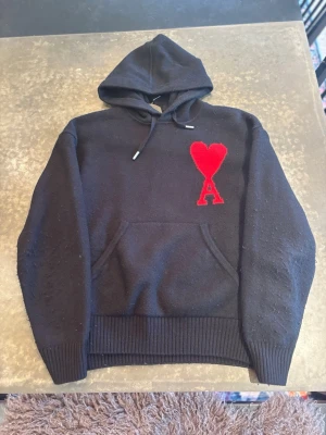 Svart hoodie från Ami Paris med röd logga - Svart hoodie från Ami Paris med stor röd hjärtformad A-logga på bröstet. Klassisk huva med dragsko, känguruficka framtill och ribbade muddar. Tillverkad i mjukt stickat material som känns riktigt skönt mot huden. Perfekt för en chill och stilren look.