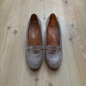 Klackskor - Snygga beige loafers från Pamroth med klack. Skorna har en elegant guldfärgad kedjedetalj framtill som ger en lyxig touch. Perfekta för dig som gillar klassisk men trendig stil.