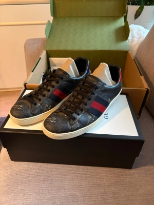 Gucci Ace sneakers med GG-mönster - Snygga Gucci Ace sneakers i svart med klassiskt GG-mönster och detaljer i rött och blått. Skorna har vita sulor, svarta skosnören och ikoniska ränder på sidan. Baksidan har metallicdetaljer i rött och blått med Gucci-logga. Perfekta för dig som vill sticka ut med lyxig streetstyle.