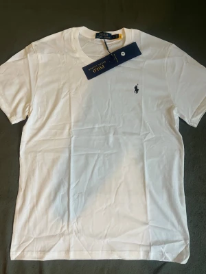Vit t-shirt från Polo Ralph Lauren - Klassisk vit t-shirt från Polo Ralph Lauren med den ikoniska lilla loggan broderad på bröstet. T-shirten har rund hals, korta ärmar och är gjord i mjuk bomull för en skön känsla. Perfekt basic-plagg till garderoben.