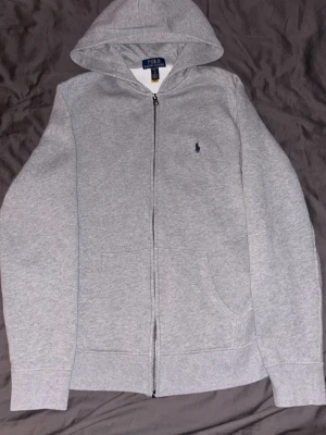 Grå hoodie från Polo Ralph Lauren - Klassisk grå hoodie med dragkedja från Polo Ralph Lauren. Hoodien har huva, två fickor framtill och den ikoniska lilla broderade loggan på bröstet. Perfekt för en avslappnad och stilren look.