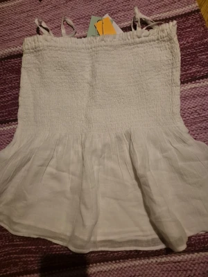 Vit linnetopp från Gina Tricot - Söt vit linnetopp från Gina Tricot med tunna axelband och smock upptill. Toppen har en volang nedtill och är tillverkad i 50% OCS-certifierad bomull. Perfekt för varma dagar och enkel att styla med jeans eller kjol.
