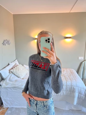 Grå stickad tröja från Zadig & Voltaire - Snygg grå stickad tröja från Zadig & Voltaire med svart och röd logotyp framtill. Tröjan har rund halsringning, långa ärmar och ribbade muddar vid ärmslut och nederkant. 