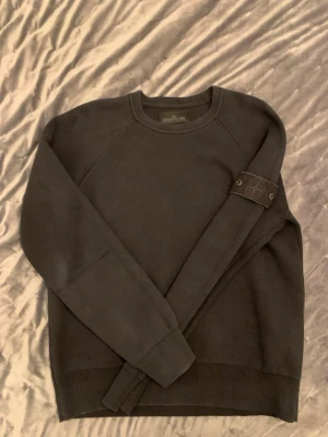 Stone Island ” ghost piece ” - Cool svart sweatshirt från Stone Island med klassisk rund hals och ribbade muddar. Ikonisk logopatch på vänster ärm som fästs med knappar. Perfekt för dig som gillar streetwear och enkel stil.