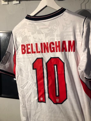 England fotbollströja Bellingham 10 - Säljer en vit England fotbollströja från Nike med BELLINGHAM och nummer 10 i rött på ryggen. Tröjan har svarta och röda detaljer, broderad landslagssköld och Nike-logga på bröstet. Perfekt för dig som vill stötta England på match eller träning.