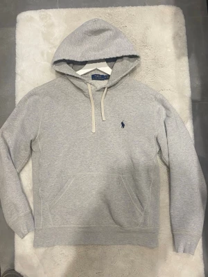 Grå hoodie från Ralph Lauren - Klassisk ljusgrå hoodie från Polo Ralph Lauren med blå broderad logga på bröstet. Hoodien har huva med vita snören och en stor magficka framtill. Tillverkad i mjuk bomullsblandning och har ribbade muddar vid ärmslut och nederkant.