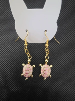 Handgjorda Guldiga örhängen med rosa sköldpaddor - Snygga handgjorda hängande örhängen i guldfärg med små sköldpaddor i rosa och guld. Sköldpaddorna har detaljerat mönster på skalet och örhängena har krokfäste. Perfekt accessoar för dig som gillar djur och vill ha något unikt.