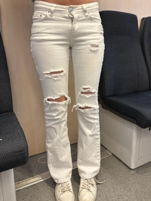 Vita ripped jeans med utsvängda ben - Säljer ett par vita jeans med utsvängda ben och flera slitningar på framsidan. Jeansen har hög midja, klassiska fem fickor och knappgylf. Materialet är denim i bomull och de har en trendig, avslappnad look.