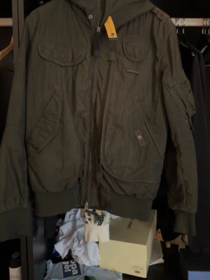 Olivgrön bomberjacka Parajumpers - Snygg olivgrön bomberjacka från Parajumpers med flera fickor, ribbade muddar och ikonisk patch på ärmen. Jackan har dragkedja framtill, huva och detaljer i orange. Perfekt för dig som gillar streetwear och funktionella plagg.