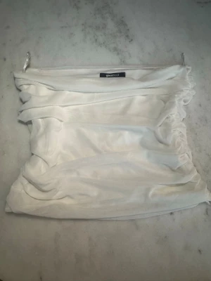 Vit bandeau tubtopp från Gina Tricot - Säljer en vit bandeau tubtopp från Gina Tricot. Toppen är ärmlös och har snygga rynkade detaljer på sidorna som ger en cool och trendig look. Tillverkad i mjukt och stretchigt material som sitter skönt på kroppen. Perfekt till sommaren!