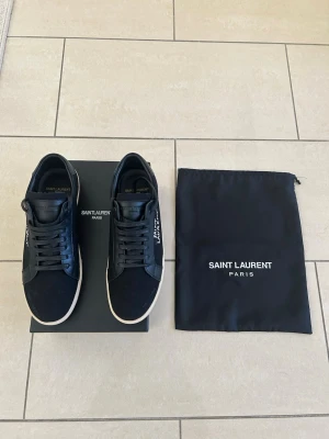 Svarta Saint Laurent sneakers - Svarta sneakers från Saint Laurent med vit sula och handskriven logga på sidan. Skorna har en klassisk låg profil, svarta skosnören och detaljer i både mocka och läder. Perfekta för dig som gillar stilrena och exklusiva sneakers. Utan box!