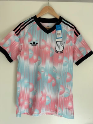 Adidas Belgien sporttröja rosa/blå - Unik sporttröja från Adidas med retroinspirerat mönster i rosa, ljusblått och vitt. Tröjan har svarta detaljer vid krage och ärmslut samt klassiska Adidas-ränder på axlarna. Tillverkad i lätt och ventilerande material, perfekt för träning eller streetwear.