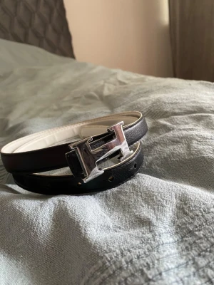 Svart skärp med H-spänne - Snyggt svart skärp med ikoniskt H-format metallspänne i silver. Bältet har en stilren design med vit insida och är tillverkat i äkta skinn. Perfekt accessoar för att lyfta vilken outfit som helst.
