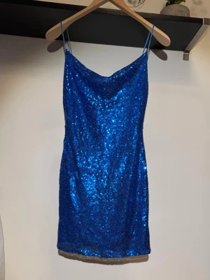 Blå glittrig paljettklänning - Snygg blå kort klänning med tunna axelband och glittriga paljetter över hela tyget. Klänningen har en lätt draperad urringning och är perfekt för dig som vill sticka ut. Perfekt för festliga tillfällen och när du vill glänsa extra.