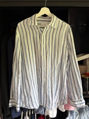 Randig linneskjorta från Massimo Dutti - Snygg vit skjorta i 100% linne från Massimo Dutti med blåa vertikala ränder. Skjortan har klassisk krage, knäppning framtill och långa ärmar med knapp vid ärmslut. Perfekt för en fräsch och avslappnad stil.