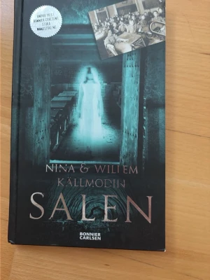 Salen av Nina & Willem Källmodin - En spännande och mystisk ungdomsroman med svart hård pärm. Omslaget har en övernaturlig känsla med en suddig figur och gamla skolbänkar. Boken handlar om vänner som upptäcker en hemlig dörr i ett skolbibliotek och möter något övernaturligt.