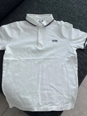 Vit pikétröja från BOSS - Klassisk vit pikétröja från BOSS med korta ärmar och krage med svarta detaljer. Tröjan har knappar framtill och BOSS-logga broderad på bröstet. Perfekt till en clean och stilren look.