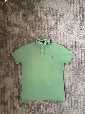 Grön pikétröja från Ralph Lauren - Klassisk grön pikétröja från Ralph Lauren med korta ärmar och krage. Tröjan har knappar framtill och den ikoniska broderade loggan på bröstet. Perfekt till jeans eller shorts för en avslappnad men snygg look.