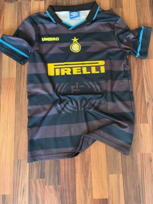 Inter Milan 90-talet retro tröja ( R9 ) - Retro Inter Milan matchtröja från Umbro med Ronaldo och nummer 10 på ryggen. Svart och mörkblåa ränder, gula detaljer och Pirelli-logga på bröstet. Klassisk krage med blå kant och korta ärmar med blåa detaljer. Perfekt för fotbollsfans!