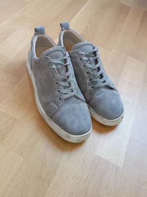 Gråa sneakers från Christian Louboutin - Snygga gråa sneakers i mocka från Christian Louboutin med klassisk röd sula och vita kanter. Skorna har lågt skaft, matchande gråa skosnören och en stilren design som passar till det mesta. Perfekta för dig som gillar exklusiva detaljer och streetstyle.