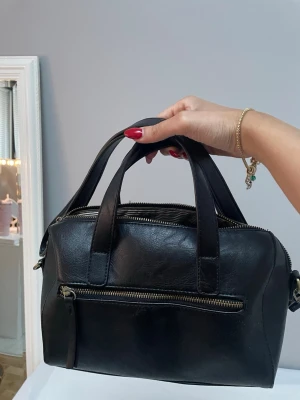 Svart väska vintage - Snygg svart handväska i skinnimitation med både handtag och avtagbar axelrem. Väskan har en praktisk dragkedjeficka framtill och stängs med dragkedja upptill. Varan är i mycket gott skick då den knappt kommit till användning, enda defekten är att dragkedjan ej fungerar. För fler frågor eller bilder är det bara att skiva! 💕