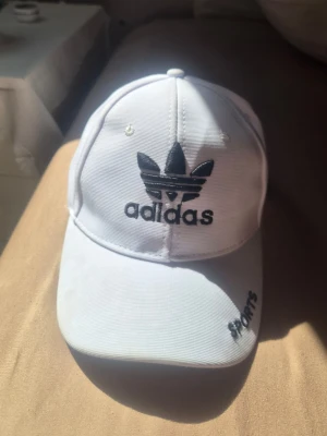 Vit keps från Adidas med brodyr - Snygg vit keps från Adidas med svart broderad logga framtill och texten 'SPORTS' på skärmen. Klassisk böjd skärm och justerbar baksida. Tillverkad i bomullsmaterial som andas. Perfekt för en sportig och trendig look.