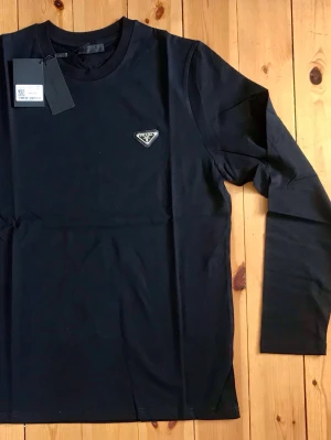 Svart långärmad topp från Prada - Snygg svart långärmad topp från Prada med rund halsringning och ikonisk logga på bröstet. Tillverkad i mjuk bomull som känns skön mot huden. Perfekt för dig som gillar stilrena och exklusiva plagg.