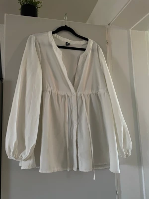 Vit blus med knappar från SHEIN - Snygg vit blus från SHEIN med knappar framtill och ballongärmar. Blusen har en avslappnad passform, rundad halsringning och dragsko i midjan. Perfekt att styla med jeans eller kjol för en trendig look.