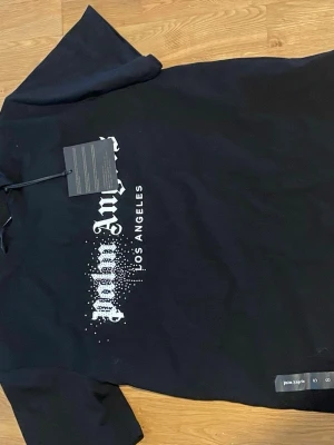 Svart Palm Angels t-shirt Los Angeles - Snygg svart t-shirt från Palm Angels med Los Angeles-tryck i vitt framtill. Klassisk passform med korta ärmar och rund hals. Tillverkad i mjuk bomull och har coolt tryck med prickdetaljer. Perfekt för dig som gillar streetwear och vill sticka ut.