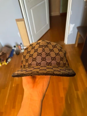 Gucci beige GG-mönstrad keps - Säljer en beige keps från Gucci med det klassiska GG-monogrammet över hela kepsen. Kepsen har böjd skärm och justerbart band bak i svart läder. Materialet är canvas med läderdetaljer. Perfekt för dig som vill ha en stilren och lyxig accessoar.