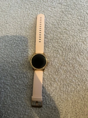 Rosa smartklocka med gulddetaljer - Snygg smartklocka med rund urtavla, guldfärgad boett och ljusrosa silikonarmband. Klockan har en slät, svart display och detaljer runt urtavlan som ger en exklusiv känsla. Perfekt för dig som vill ha koll på både hälsa och notiser direkt på handleden.