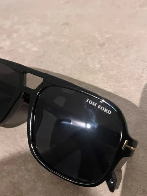 Svarta Tom Ford solglasögon - Snygga svarta solglasögon från Tom Ford med klassisk pilotform och mörka glas. Bågarna har gulddetaljer vid gångjärnen och en cool design med en liten öppning vid näsryggen. Perfekta för dig som vill ha en stilren och trendig look.