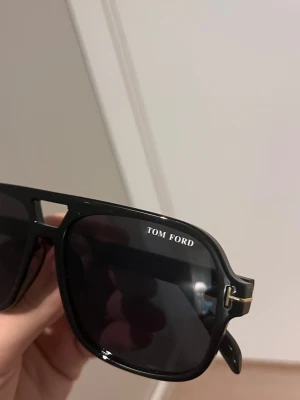 Svarta Tom Ford solglasögon - Snygga svarta solglasögon från Tom Ford med klassisk och stilren design. De har en fyrkantig form, mörka linser och diskreta gulddetaljer på bågarna. Perfekta för att höja din look under soliga dagar.