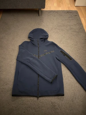Mörkblå Nike hoodie med dragkedja - Snygg mörkblå hoodie från Nike med hel dragkedja framtill och huva. Hoodien har en diskret Nike-logga på bröstet och en svart rand över bröstet. Ficka med dragkedja på ena ärmen och ribbade muddar. Perfekt för en sportig och avslappnad stil.