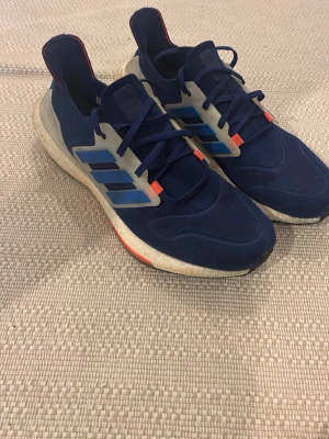 Adidas ultra boost blå - Snygga blå sneakers från Adidas med klassiska tre ränder i ljusblått på sidan. Ovandelen är i mesh och syntet, och sulan är en vit boost-sula med röda detaljer. Skorna har lågt skaft och sportig design, perfekta för en aktiv stil.