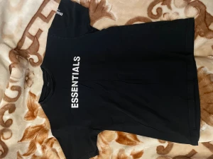 Svart Essentials t-shirt - Snygg svart t-shirt från Essentials med vit text på bröstet. Klassisk rak passform och rund halsringning. Tillverkad i mjukt bomullsmaterial som känns skönt mot huden. Perfekt för en clean och stilren look. Essential märket på framsidan av tröjan är lite skrynkliga och är inte i de bästa skicket men annars är hela tröjan väldigt bra skick.