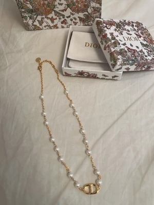halsband med pärlor och guld - Lyxigt halsband med guldfärgad kedja och vita pärlor längs hela halsbandet. Mittendelen har en ikonisk CD-logga i guld. Kommer i original ask med blommigt mönster. Perfekt för dig som vill ha något elegant och trendigt. Utan paketering och box är det 350kr men med blir det 550kr