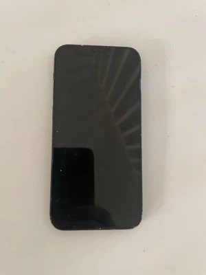 Svart iPhone 12 med dubbla kameror - Säljer en svart iPhone med stilren design och rundade hörn. Telefonen har dubbla kameror på baksidan och den klassiska Apple-loggan. Tillverkad i glas och metall, perfekt för dig som gillar snygg och modern teknik.