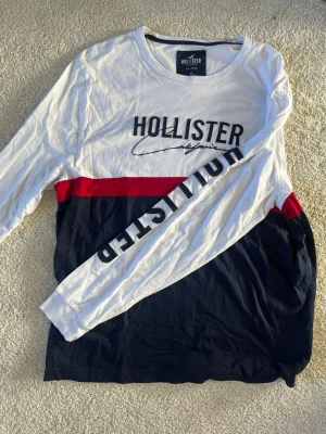 Långärmad t-shirt från Hollister - Långärmad t-shirt från Hollister i bomull med vit överdel, svart nederdel och en röd rand över bröstet. Stor Hollister-logga tryckt på bröstet och längs ena ärmen för en sportig vibe.