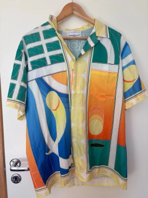 Färgstark silkesskjorta  - Unik kortärmad skjorta från Casablanca i silke med färgstarkt grafiskt mönster i grönt, blått, orange, gult och vitt. Skjortan har klassisk krage och knappar framtill. Perfekt för dig som vill sticka ut med en exklusiv och trendig look.