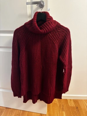 Vinröd polotröja från Amisu - Mysig vinröd stickad polotröja från Ambiguous med grov ribbstickning och hög krage. Tröjan har lång ärm och avslappnad passform, perfekt för kalla dagar. Materialet är mjukt och värmande, vilket gör den extra skön att bära.