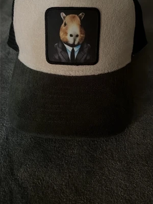 Beige och svart keps med capybara - Säljer en beige och svart keps med mesh baktill och en unik patch framtill som visar en capybara i kostym. Kepsen har böjd skärm och justerbar snapback. Perfekt för dig som gillar djurmotiv och vill sticka ut med din stil.